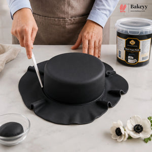 Person cutting a black fondant cake with a knife on a white surface, featuring Bakeyy branding.