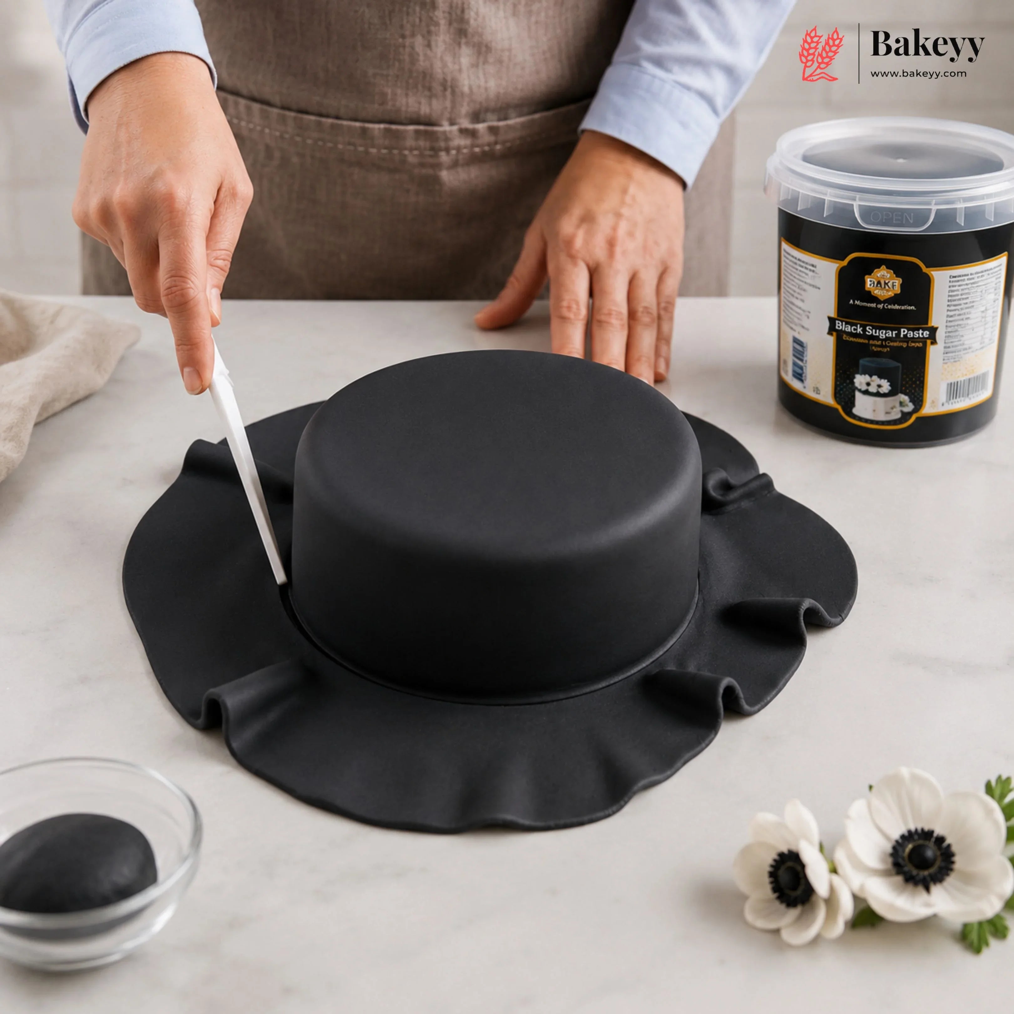 Person cutting a black fondant cake with a knife on a white surface, featuring Bakeyy branding.