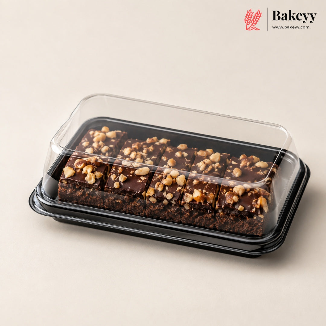 Chocolate nut bars in a clear plastic clamshell container with a black tray, photographed on a warm beige background. The Bakeyy logo is visible in the top right corner.