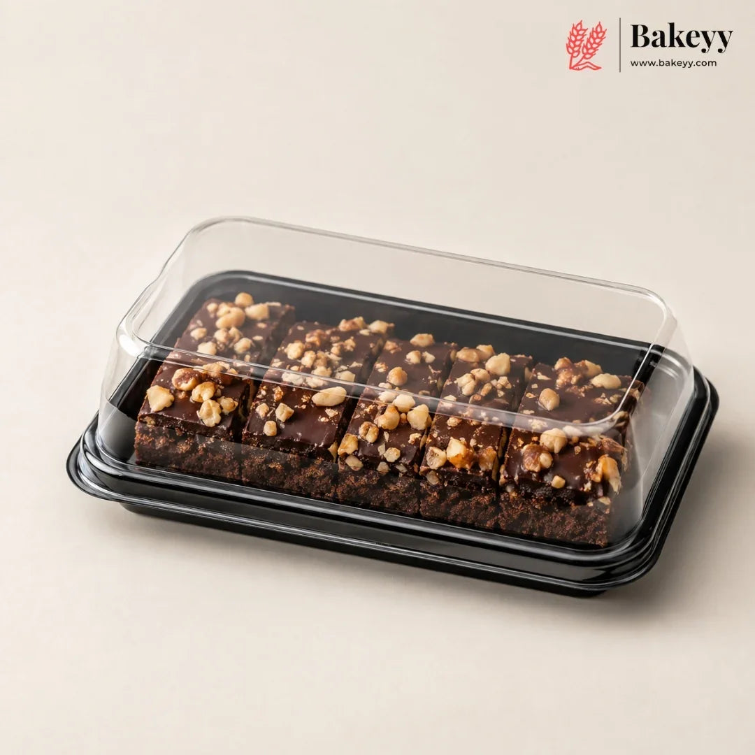 Chocolate nut bars in a clear plastic clamshell container with a black tray, photographed on a warm beige background. The Bakeyy logo is visible in the top right corner.