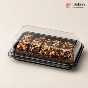 Chocolate nut bars in a clear plastic clamshell container with a black tray, photographed on a warm beige background. The Bakeyy logo is visible in the top right corner.