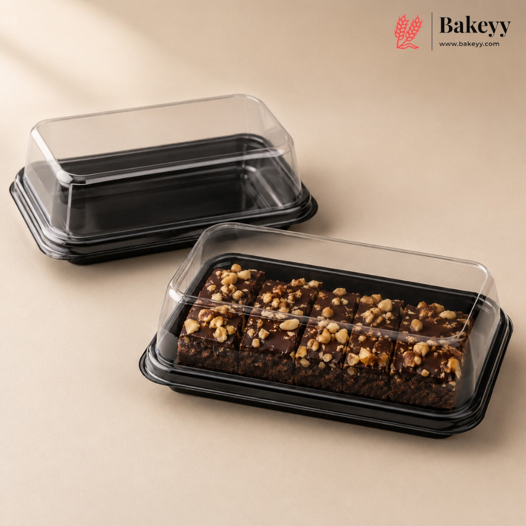 Two clear plastic bakery boxes with black bases are shown on a warm beige background. The front box contains chocolate bars topped with chopped nuts, while the rear empty box sits behind it.
