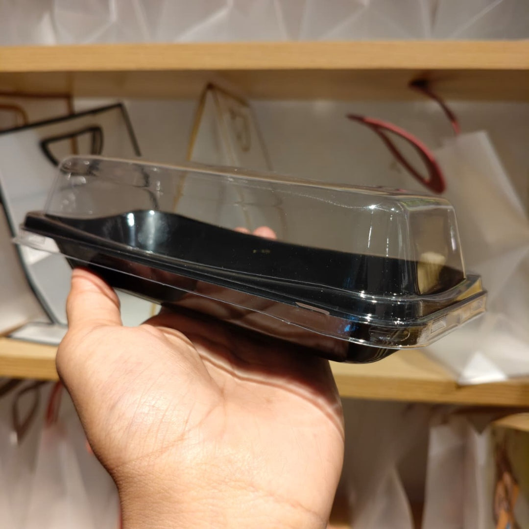 A hand holding a clear plastic clamshell container with a black tray insert, shown in front of a wooden shelf with white bags in the background.