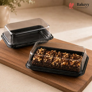 Two black plastic bakery boxes with clear lids, one empty and one holding chocolate bars topped with nuts, displayed on a wooden board with a Bakeyy logo in the corner.