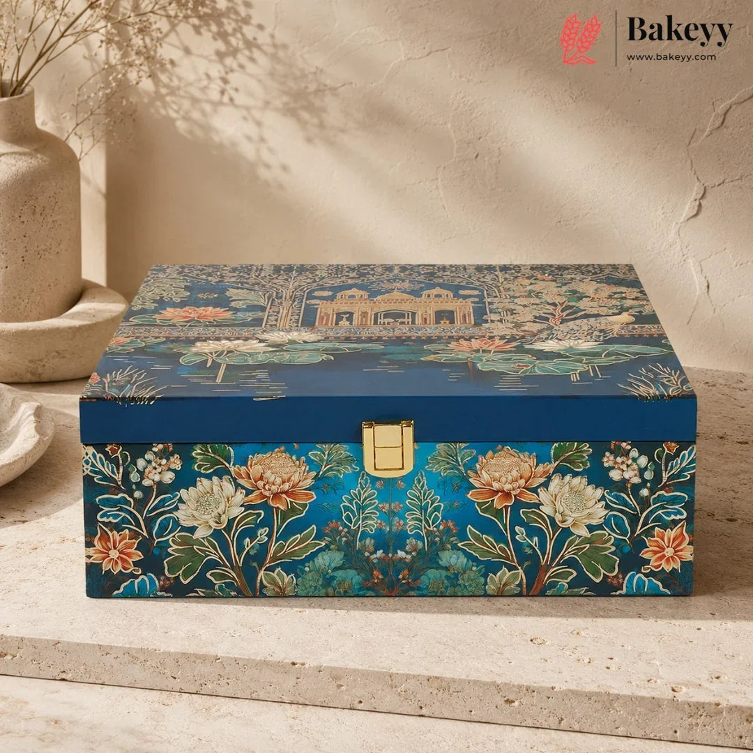 A decorative rectangular storage box with a blue lid featuring a garden pond scene and a floral front panel, shown on a stone surface against a textured beige wall. The Bakeyy logo appears in the top right corner.