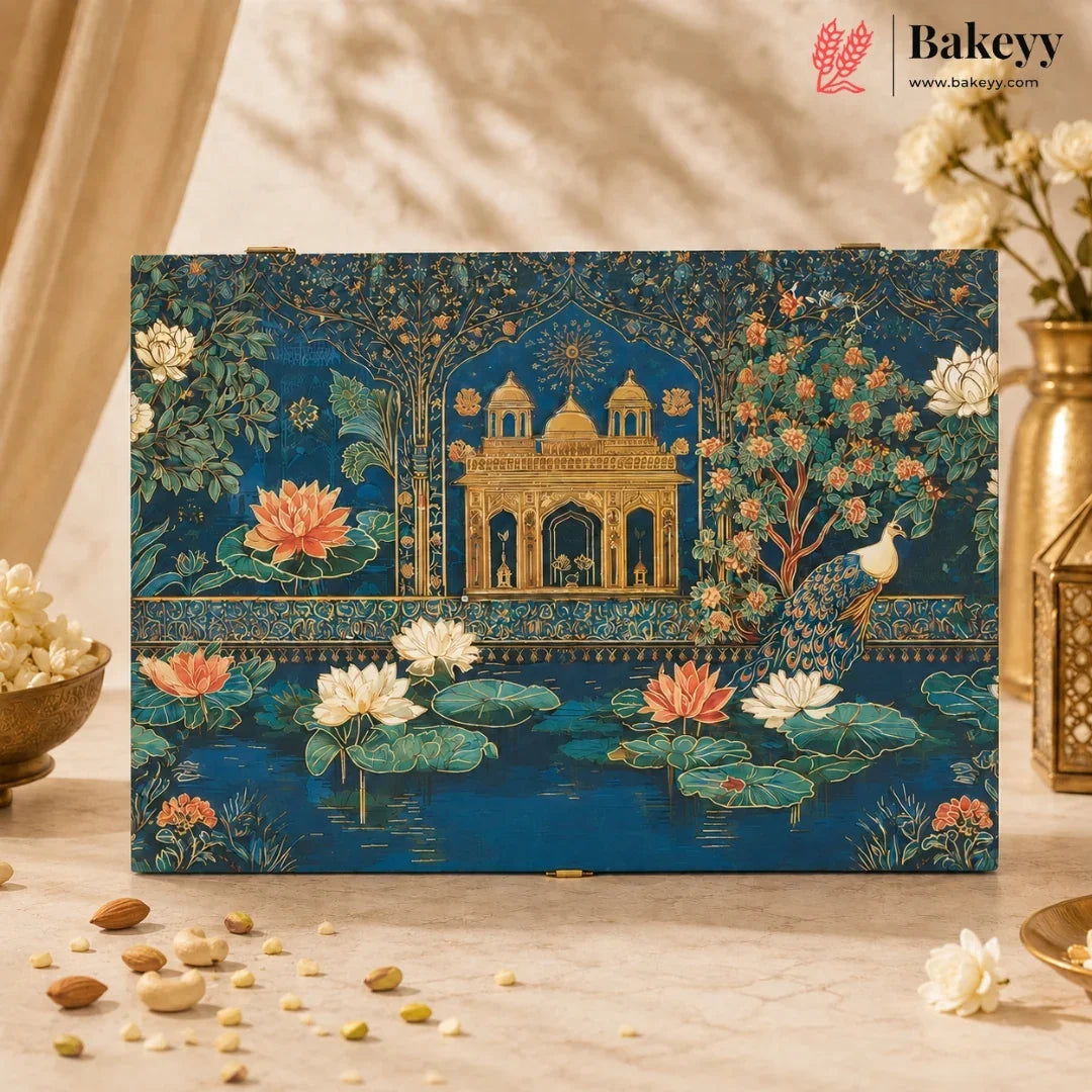 A rectangular decorative box with an ornate blue and gold illustration of a palace, peacock, lotus flowers, and trees, displayed on a beige surface with scattered nuts and flowers around it. The Bakeyy brand logo appears in the top right corner.