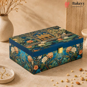 A rectangular decorative gift box by Bakeyy, featuring an intricate classical architectural and floral pattern in deep teal, gold, and orange. The box has a gold-tone metal clasp and is styled on a beige stone surface with a ceramic vase and scattered almonds nearby.