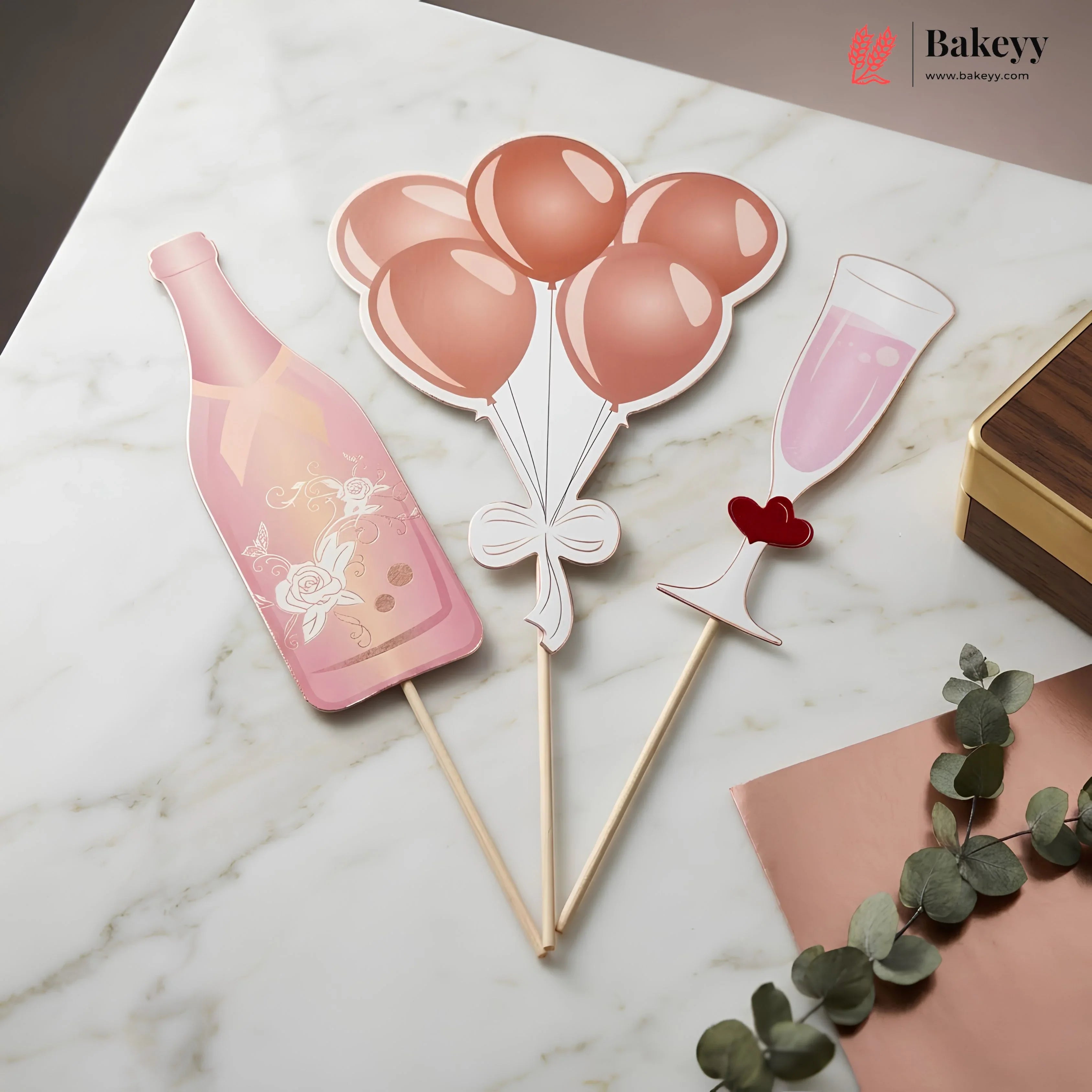 Decorative cake toppers shaped like a bottle, balloons, and a glass on a marble surface with Bakeyy branding.