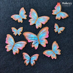 Seven butterfly-shaped sugar cookies with iridescent pastel wings in shades of blue, pink, and purple, arranged on a dark textured background. The Bakeyy logo is visible in the top right corner.