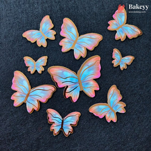Seven butterfly-shaped sugar cookies with iridescent pastel wings in shades of blue, pink, and purple, arranged on a dark textured background. The Bakeyy logo is visible in the top right corner.