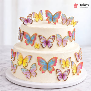 A two-tier white frosted cake decorated with colorful butterfly toppers in pink, yellow, blue, and purple, displayed on a white cake stand against a bright white background. The Bakeyy logo is visible in the top right corner.