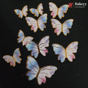 Ten iridescent butterfly-shaped cake toppers with gold edges, displayed on a dark textured background. The toppers feature a pastel gradient of blue, pink, and purple with gold outlines.