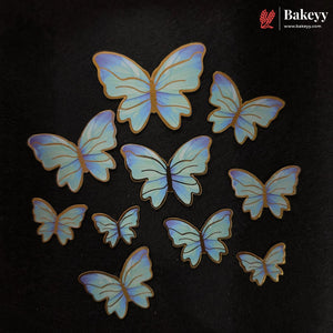 Nine iridescent blue and purple butterfly-shaped cake toppers with gold edges, arranged on a black background. The Bakeyy brand logo is visible in the top right corner.