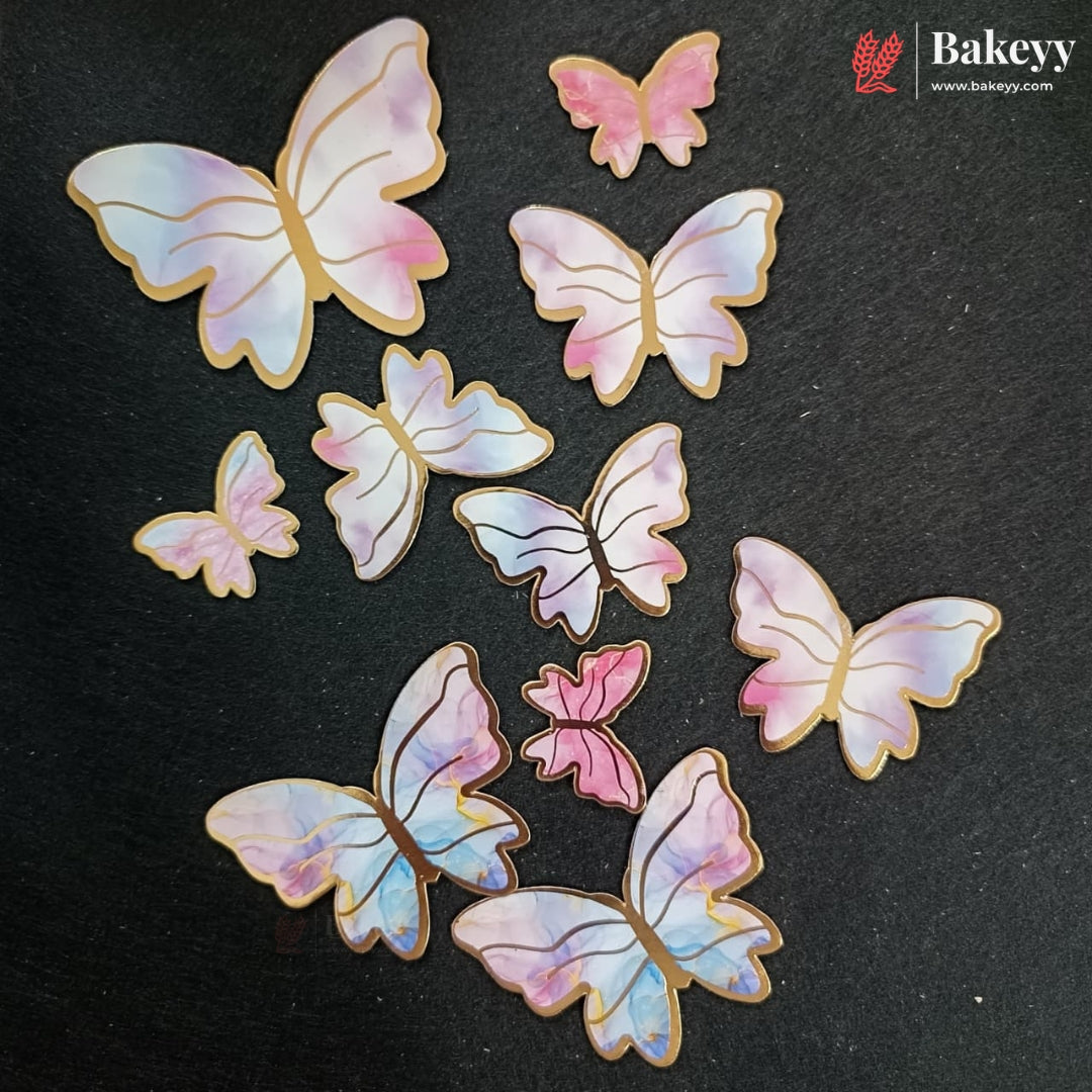 A set of decorative butterfly-shaped cookies with iridescent pastel wings in pink, blue, and white, outlined with a golden-brown border, arranged on a dark textured background. A Bakeyy logo is visible in the top right corner.