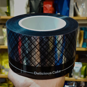 A hand holds a roll of black tape printed with a diamond grid pattern and the text 'Delicious Cake' on a blurred store shelf background.