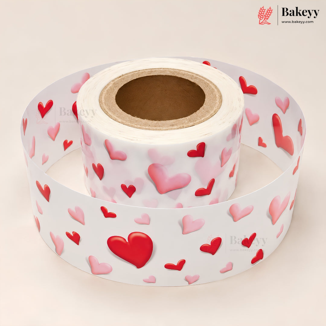 A roll of white adhesive labels printed with a pattern of red and pink 3D-effect hearts, shown with one end unrolled. The Bakeyy brand logo is visible in the top right corner.