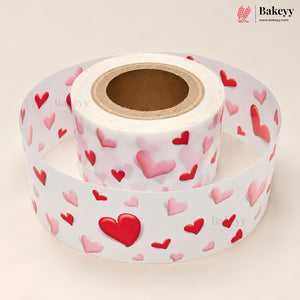 A roll of white adhesive labels printed with a pattern of red and pink 3D-effect hearts, shown with one end unrolled. The Bakeyy brand logo is visible in the top right corner.