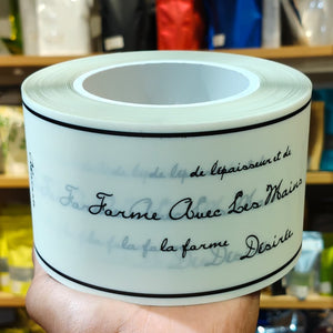 A hand holds a roll of white labels printed with elegant black cursive French text, set against a blurred background of shelves with colorful items.