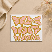 Sheet of pink and gold butterfly stickers on a beige background with a textured fabric and dried flowers.