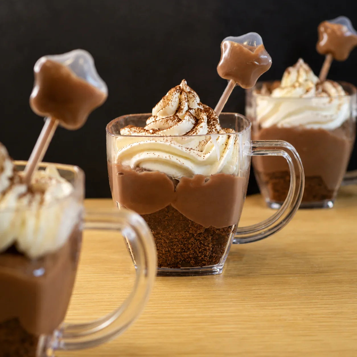 Mugs of hot chocolate with whipped cream and marshmallow sticks on a wooden surface.