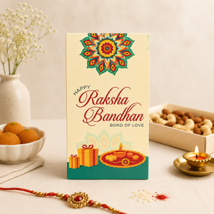 A rectangular greeting card standing upright on a tabletop, printed with 'Happy Raksha Bandhan Bond of Love' and a colorful mandala design. The card is styled in a warm home setting with a red and gold Rakhi in the foreground, a lit brass diya, a bowl of orange fruits, and a box of nuts in the background.