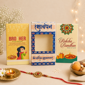 Three decorative Rakhi Bandhan gift boxes arranged on a light beige surface with a red thread and a gold-toned ornament in front. The left box is yellow with red text reading 'BROTHER WE ARE INDEEDLY'; the center box is blue and white with a window cutout and Hindi text; the right box is cream with a colorful mandala and 'Happy Rakhi Bandhan' text.