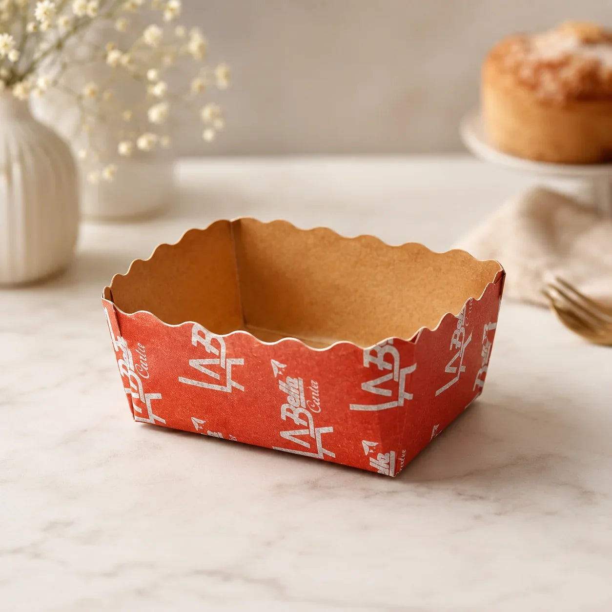 Red paper dessert box with white text on a marble surface, with a blurred background of a cake and flowers.
