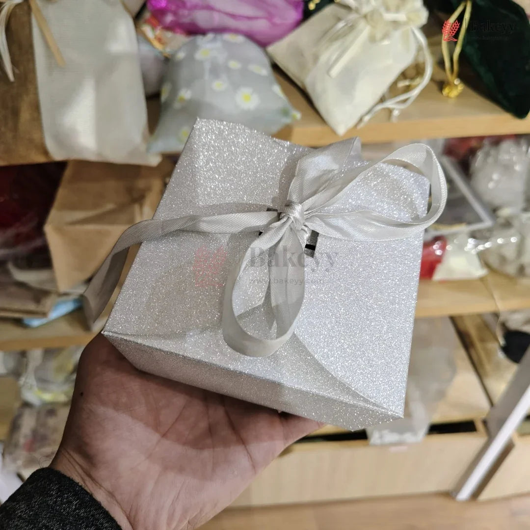 Silver glittery gift box with a bow held in a hand against a blurred background of wrapped gifts.