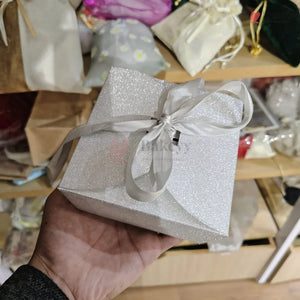 Silver glittery gift box with a bow held in a hand against a blurred background of wrapped gifts.