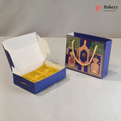 A blue and gold bakery gift box shown open with a white interior and gold tray insert, alongside a matching blue gift bag with gold rope handles and decorative gold artwork. The photo is on a light neutral studio surface with a gray backdrop.