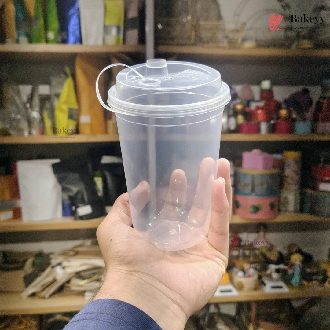 Hand holding a clear plastic cup with a lid against a blurred background of shelves with various items.