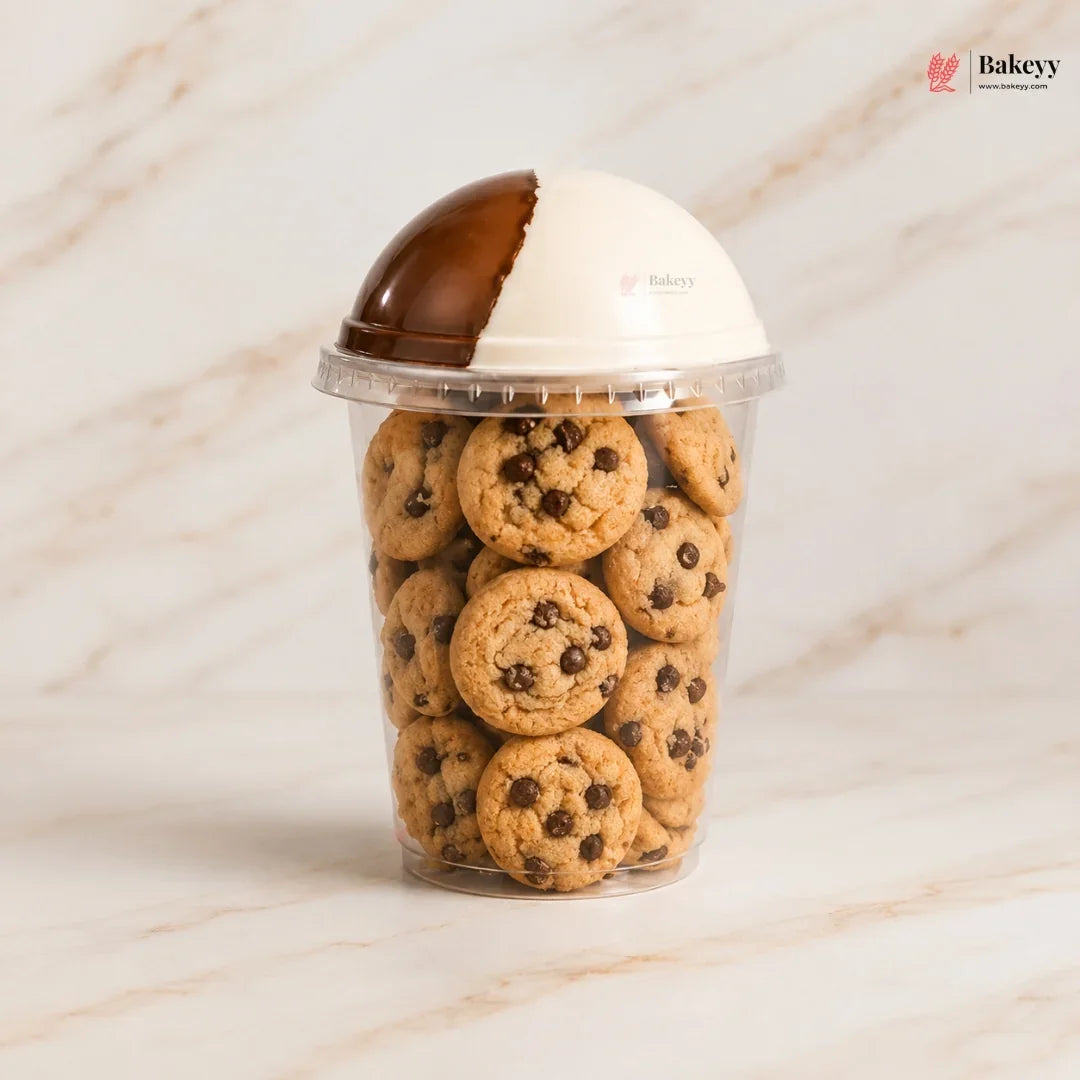 A clear plastic cup filled with chocolate chip cookies, topped with a dome-shaped lid that is split into brown and white halves, set on a light marble surface.