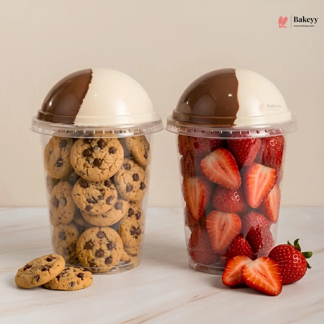 Two clear plastic deli cups with dome-shaped lids, one filled with chocolate chip cookies and the other with sliced strawberries. The lids are decorated with a diagonal split of white and dark brown chocolate.