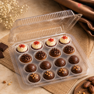 A clear plastic tray holding 12 assorted chocolate treats: four cream swirls topped with red crumbs, four plain milk-chocolate truffles, four dark-chocolate truffles, and four chocolate cupcakes with caramel and nuts. The tray is photographed on a beige fabric surface with decorative dried flowers, chocolate pieces, and draped fabric in the background.