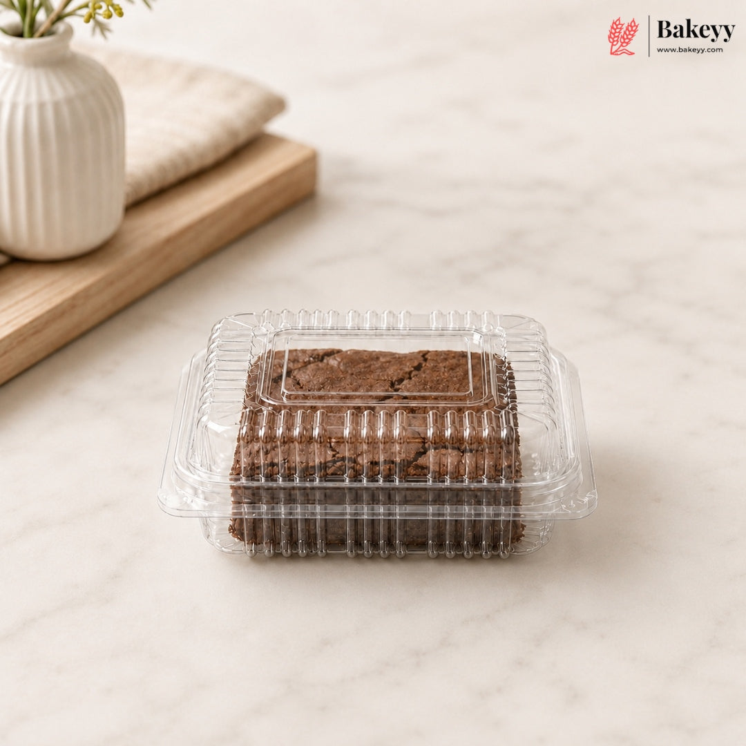 A clear plastic clamshell container holding two chocolate brownies, placed on a light marble surface with a white vase and wooden board in the background. The Bakeyy logo appears in the top right corner.