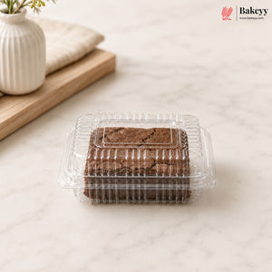 A clear plastic clamshell container holding two chocolate brownies, placed on a light marble surface with a white vase and wooden board in the background. The Bakeyy logo appears in the top right corner.