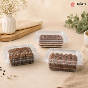 Three clear plastic clamshell boxes containing chocolate-layered cakes or pastries, arranged on a beige fabric surface with decorative props including a white vase with dried flowers, green leaves, and a small cup. The Bakeyy logo appears in the top right corner.