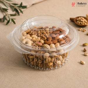 A clear plastic round container with a matching lid, filled with assorted nuts including almonds, cashews, and pistachios, photographed on a beige surface with small decorative props and a Bakeyy logo in the top right.