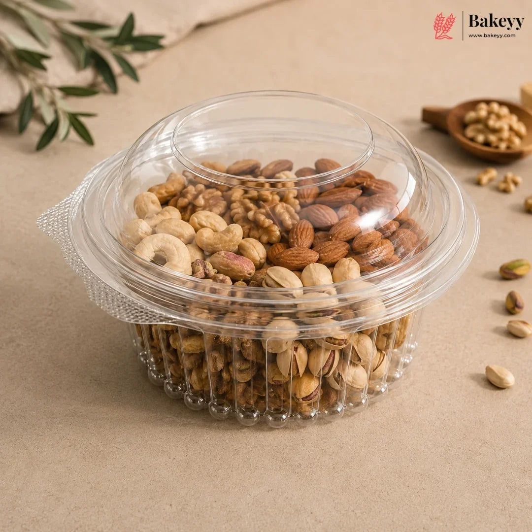 A clear plastic round container with a matching lid, filled with assorted nuts including almonds, cashews, and pistachios, photographed on a beige surface with small decorative props and a Bakeyy logo in the top right.