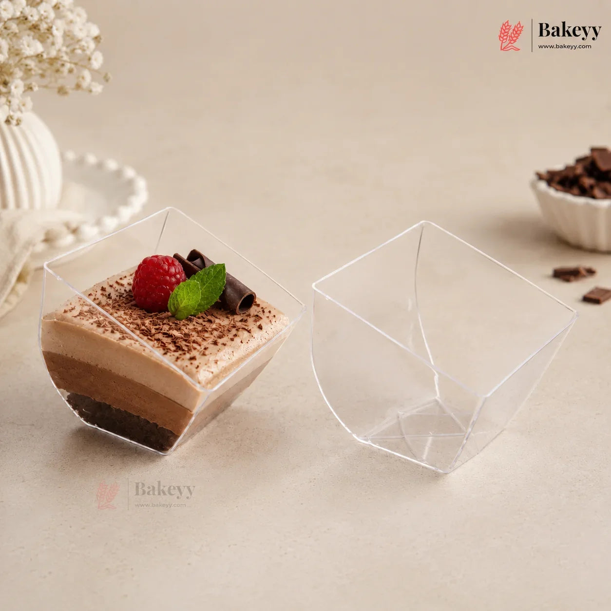 Two clear plastic dessert cups on a beige surface, with one cup filled with a layered chocolate dessert topped with a raspberry and mint, and the other cup shown empty.