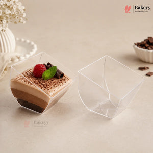 Two clear plastic dessert cups on a beige surface, with one cup filled with a layered chocolate dessert topped with a raspberry and mint, and the other cup shown empty.