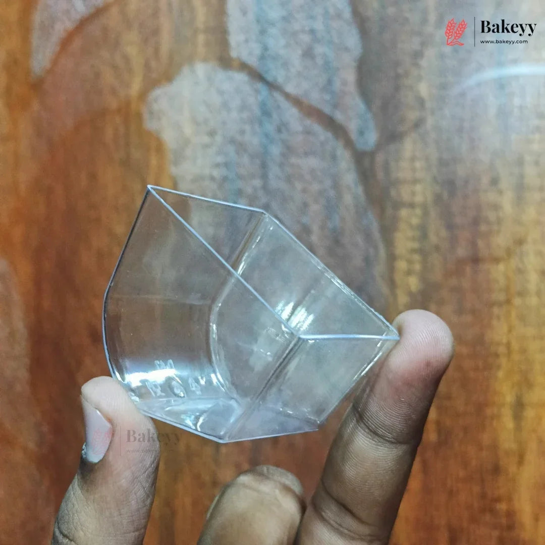 A clear plastic cup with a geometric, faceted design held by a person's fingers against a wooden background. A 'Bakeyy' brand logo is visible in the top right corner.