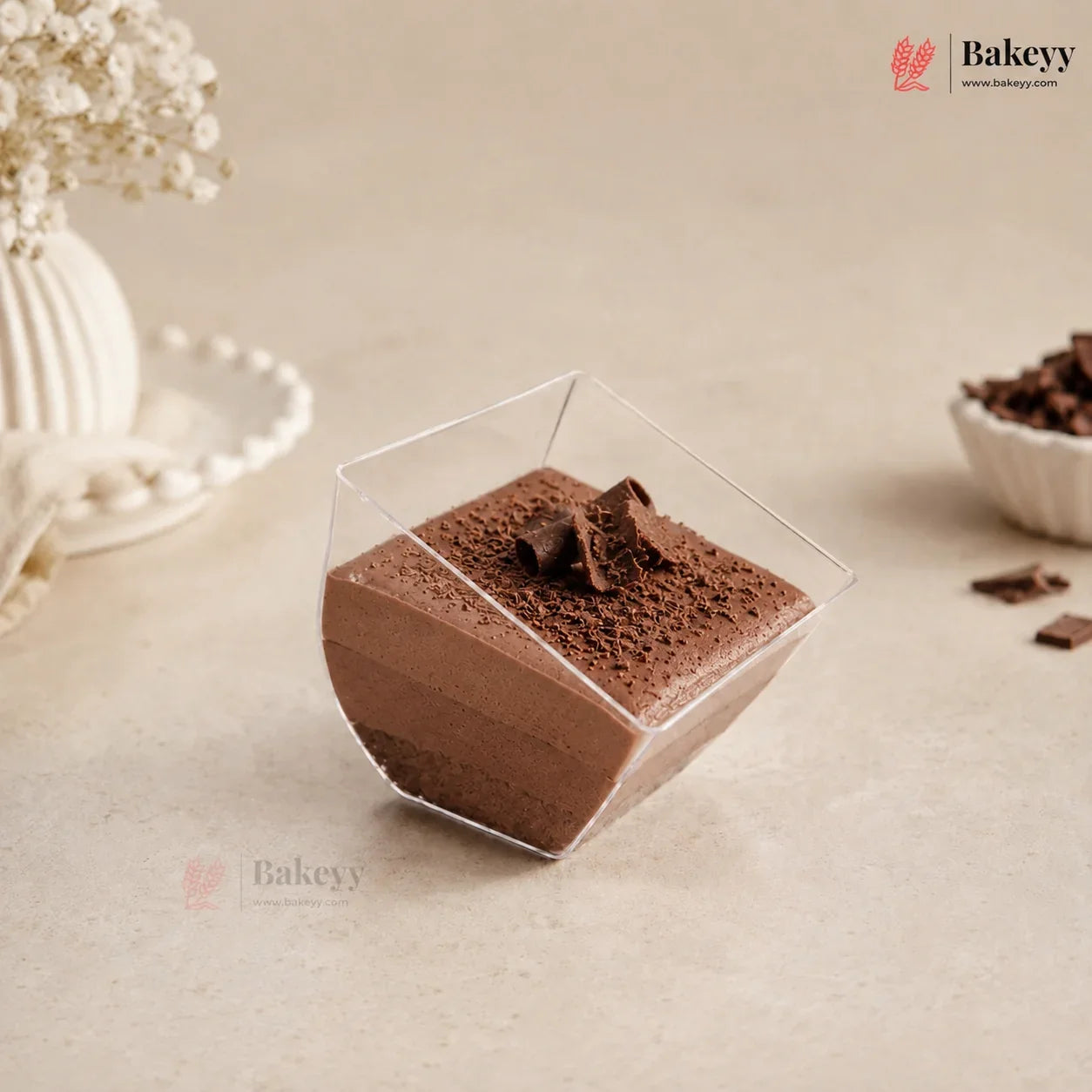 A square clear plastic dessert cup filled with chocolate mousse, topped with chocolate shavings, set on a beige surface with decorative dried flowers and a small bowl of chocolate pieces in the background.