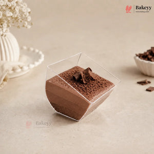 A square clear plastic dessert cup filled with chocolate mousse, topped with chocolate shavings, set on a beige surface with decorative dried flowers and a small bowl of chocolate pieces in the background.