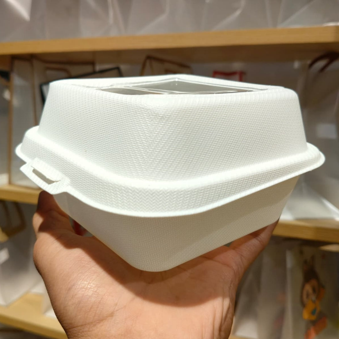 A hand holding a white textured disposable plastic food container with a clear plastic window on the lid, set against a blurred background of wooden shelves and white bags.