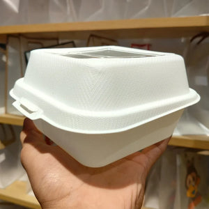 A hand holding a white textured disposable plastic food container with a clear plastic window on the lid, set against a blurred background of wooden shelves and white bags.