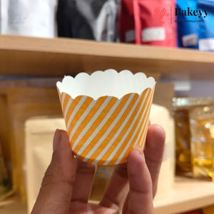 A hand holding a paper cup liner with orange and white diagonal stripes, with a bakery shelf blurred in the background. The Bakeyy logo is visible in the top right corner.