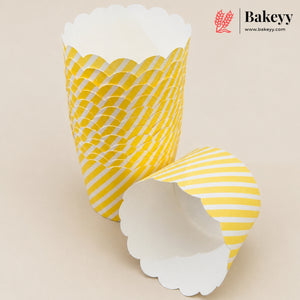 Three yellow and white patterned paper cupcake liners with scalloped edges, shown on a light beige background. One liner is standing upright, showing a yellow and white wavy pattern, while another is lying on its side showing a yellow and white diagonal stripe pattern.