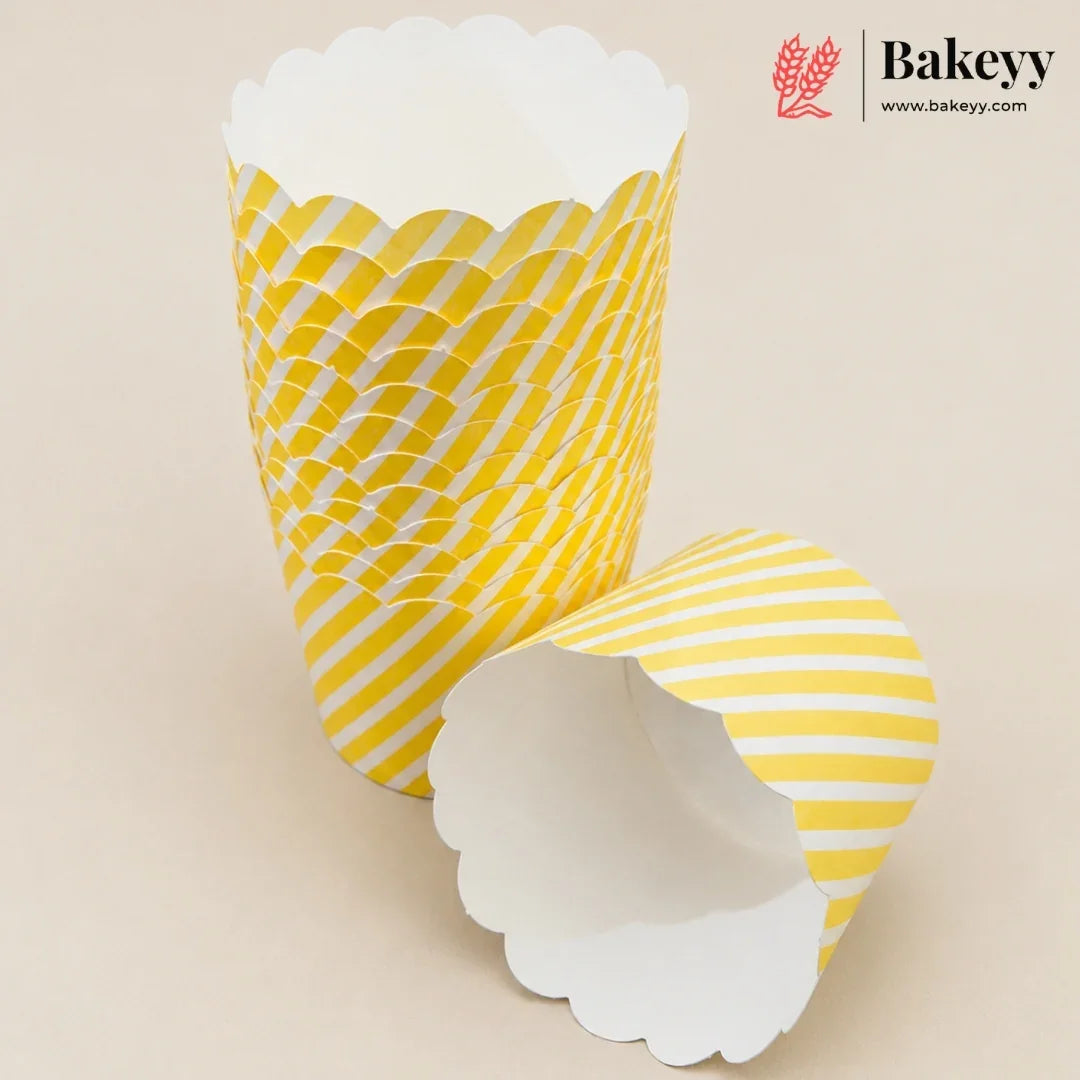 Three yellow and white patterned paper cupcake liners with scalloped edges, shown on a light beige background. One liner is standing upright, showing a yellow and white wavy pattern, while another is lying on its side showing a yellow and white diagonal stripe pattern.