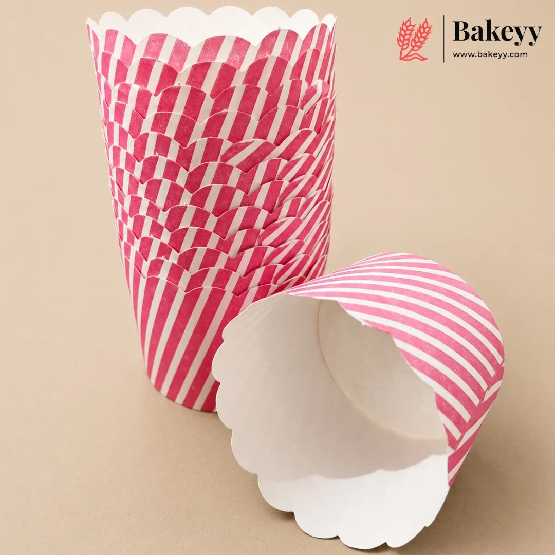 A stack of pink and white striped scalloped paper cupcake liners with one liner lying open in the foreground on a beige background. The Bakeyy logo is visible in the top right corner.